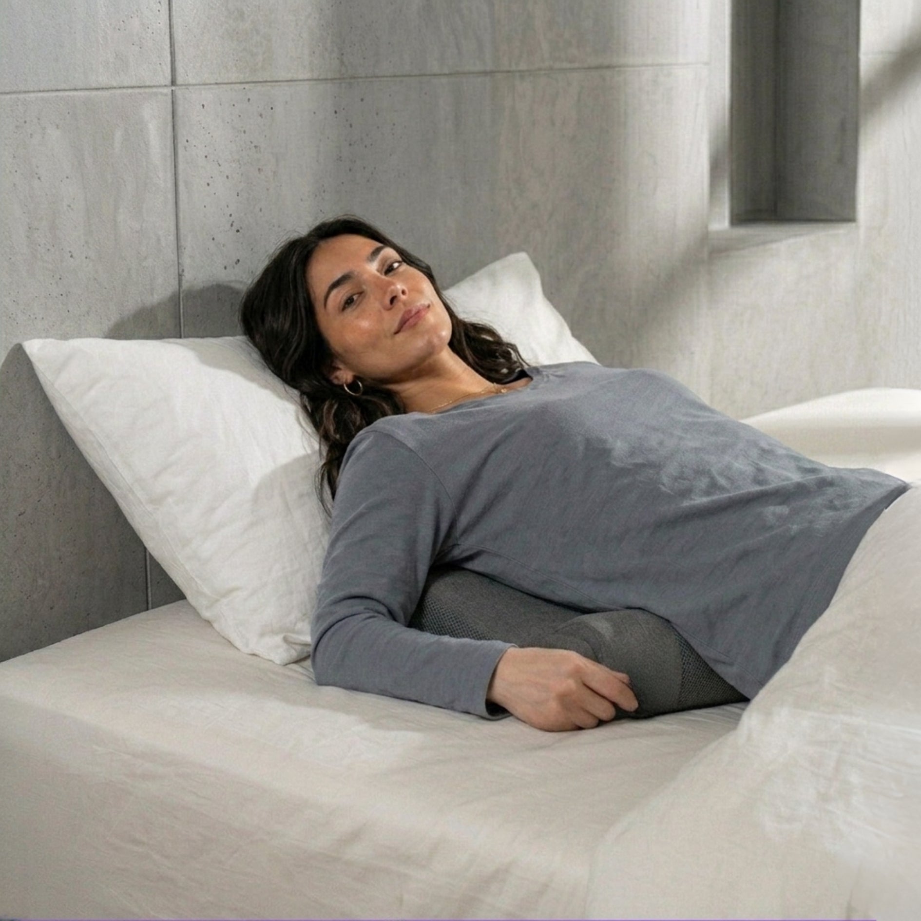 Woman lying on a bed with the Threvek Pulse, wearing a gray long-sleeve shirt and dark pants.